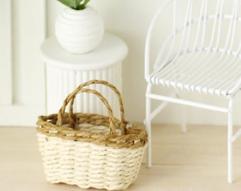 Puede incluir: Una pequeña cesta tejida con dos asas. La cesta está hecha de materiales beige claro y marrón. La cesta está sobre una superficie de madera clara. Una pequeña mesa blanca con una planta y una silla blanca están en el fondo.
