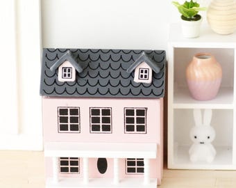 May include: A pink and gray wooden dollhouse with a white porch and black windows. The roof is gray with a scalloped design.
