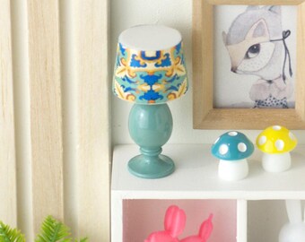 May include: A miniature teal lamp with a patterned lampshade featuring blue, yellow, and white designs. The lamp sits on a white shelf with a framed picture of a squirrel, two mushroom figurines, and a pink balloon dog.