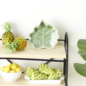 May include: A miniature display featuring a green leaf-shaped dish, two small pineapples, a bowl of yellow fruit, and a tray of green grapes. The items are arranged on wooden shelves with a black metal frame, creating a charming, decorative scene.