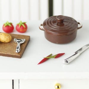 Può includere: Un set da cucina in miniatura con una pentola in ceramica marrone, un tagliere di legno, uno spelucchino per patate argentato, due pomodori rossi, una patata e due peperoncini rossi.