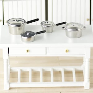 May include: A set of four miniature silver pots with black handles on a white table with two drawers and a shelf. The pots have glass lids.