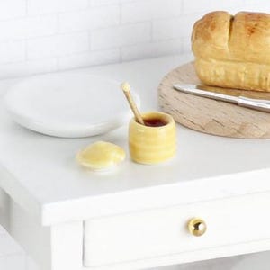 May include: A miniature honey pot with a wooden dipper, a small white plate, and a loaf of bread on a wooden chopping board. The honey pot is yellow and white with a brown lid. The dipper is brown and has a white tip. The plate is white and round. The bread is white and has a crust. The chopping board is brown and rectangular.
