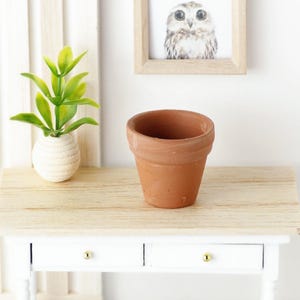 Puede incluir: Una maceta de terracota sobre una mesa blanca con una tapa de madera. La maceta está vacía y tiene una textura rugosa.