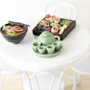 Könnte beinhalten: Ein Miniatur-Tischset in Weiß mit einer grünen Teekanne und Teetassen, einer Schüssel Ramen und einem Tablett mit Sushi. Der Tisch ist von weißen Stühlen umgeben.