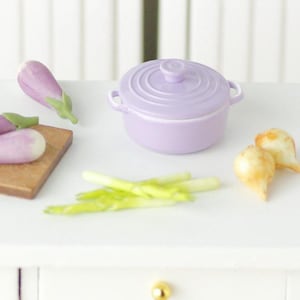 Puede incluir: Una olla de cerámica miniatura de color púrpura con tapa, una tabla de cortar de madera con berenjenas moradas, espárragos verdes y pequeñas cebollas marrones.