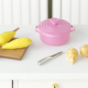 Puede incluir: Una olla de cerámica rosa en miniatura con tapa, dos mazorcas de maíz amarillas sobre una tabla de cortar de madera, dos cebollas pequeñas marrones y un cuchillo plateado sobre una encimera blanca.