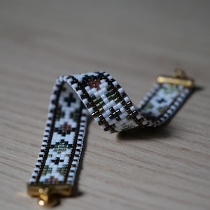 Peut inclure: Un bracelet en perles tissées présentant un motif géométrique composé de perles en verre blanc, brun foncé et vert sourd. Le bracelet est terminé par des embouts et un fermoir en métal doré, affichant un design symétrique répétitif.