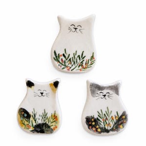 Puede incluir: Tres adornos de cerámica blanca con forma de gato que presentan caras pintadas a mano con ojos cerrados y bigotes. Cada gato muestra un patrón botánico único en la base, incluyendo hojas verdes y pequeños acentos naranjas o negros.