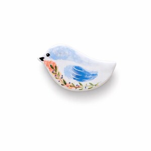 Puede incluir: Un objeto de cerámica blanca con forma de pájaro que presenta un diseño pintado a mano con cabeza y ala de color azul claro, pico negro y una guirnalda decorativa de pequeñas flores naranjas y hojas verdes en la parte inferior.