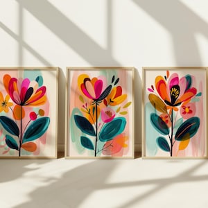 May include: Three framed floral art prints. Each print features a vibrant, abstract flower design with pink, orange, and teal hues. The flowers have dark green leaves and are set against a soft, pastel background. The frames are a light wood color.