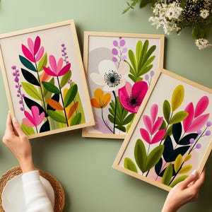 May include: Three framed floral art prints featuring colorful stylized flowers and leaves in shades of pink, green, orange, and white. The prints are held against a light green background, with a neutral-toned frame.