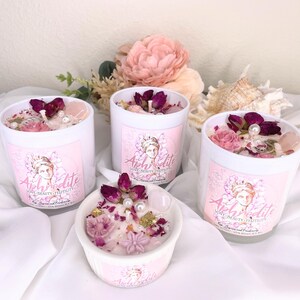 May include: Four white candles with pink labels and floral arrangements. The candles are decorated with dried rose buds, pearls, and crystals. The labels feature a portrait of a woman and the word "Aphrodite".