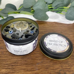 May include: A black tin candle with a gray wax interior, adorned with black stones, blue eye beads, and dried herbs. The candle's label reads "Shielded" and "Fairy Wall of Protection Candle". The lid, placed beside it, reads "Shield of Protection".