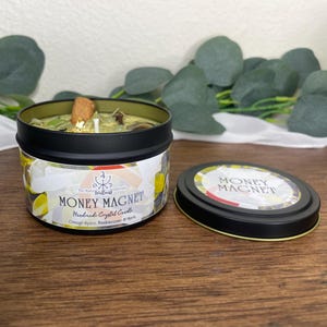 May include: A black tin candle labeled "Money Magnet" with a green wax interior. The candle includes a cinnamon stick, gold flakes, and other botanicals. The lid is next to the candle, and the background is a wooden surface.