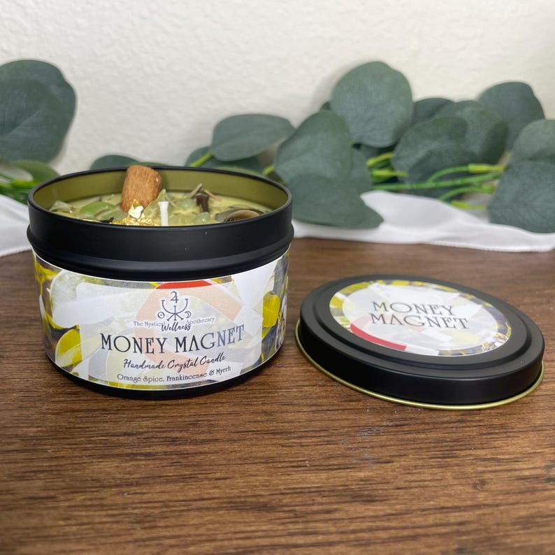 May include: A black tin candle labeled "Money Magnet" with a green wax interior. The candle includes a cinnamon stick, gold flakes, and other botanicals. The lid is next to the candle, and the background is a wooden surface.