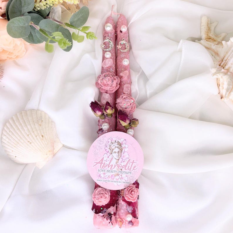 May include: Two pink candles decorated with pearls, dried roses, and floral accents. Each candle has a decorative ring near the top. A round sticker with the word "Aphrodite" and a graphic of a woman's face is at the base of the candles.