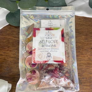 May include: A clear, iridescent package containing a 'Self Love Spiritual Bath' kit. The kit includes bath salts, rose petals, and small candles. The label reads 'Self-love, love attraction, open heart, affection.' The package is 8 oz.