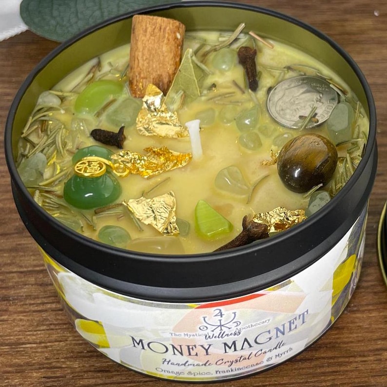 May include: A Money Magnet handmade crystal candle in a black tin. The candle has a yellow wax base with various decorative elements, including green stones, gold flakes, and a cinnamon stick. The label reads "Money Magnet" and "Orange Spice, Frankincense & Myrrh."