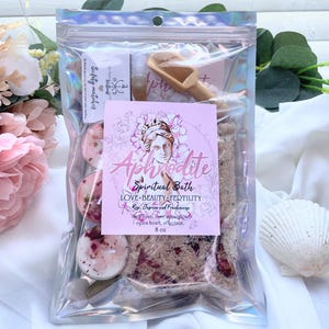 May include: A clear, iridescent bag containing a spiritual bath kit. The kit includes candles, bath salts, a wooden scoop, and a small bottle. A pink label reads "Aphrodite Spiritual Bath" with text about love, beauty, and fertility. The kit is 8 oz.