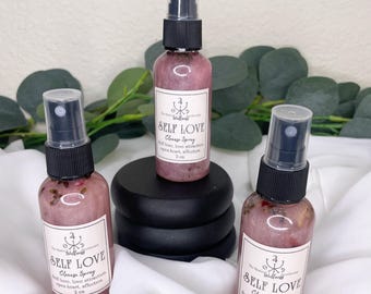 May include: Three pink Self Love Cleanse Spray bottles with black spray nozzles and white labels. The labels read "Self Love, Cleanse Spray, self-love, love attraction, opens heart, affection, 2 oz." The bottles are on a white cloth with greenery in the background.