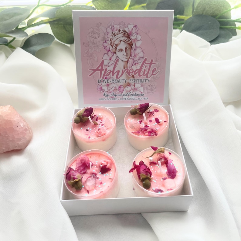 May include: A white box containing four pink candles, each adorned with dried rose buds and petals. The box lid features the word "Aphrodite" with an illustration of a woman and the words "Love, Beauty, Fertility."