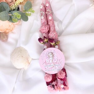 May include: Two pink intention candles decorated with pink roses, pearls, and silver charms. The candles are adorned with dried rose buds and a round label with the word "Aphrodite" and the words "Love, Beauty, Fertility".