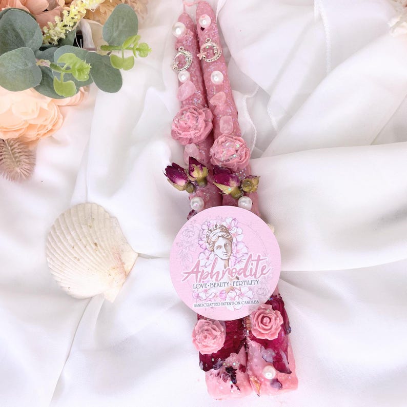 May include: Two pink intention candles decorated with pink roses, pearls, and silver charms. The candles are adorned with dried rose buds and a round label with the word "Aphrodite" and the words "Love, Beauty, Fertility".
