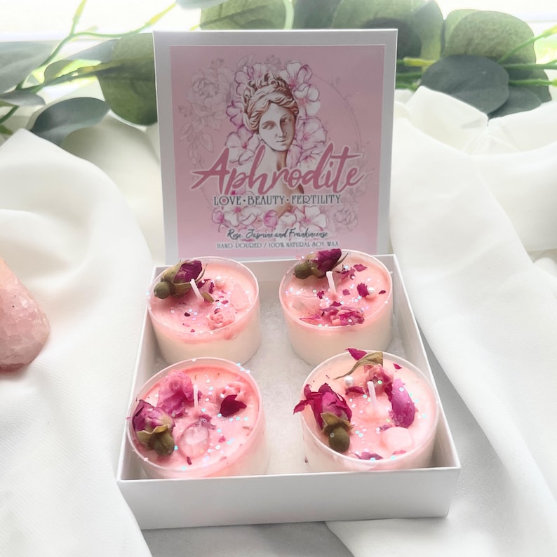 May include: A white box containing four small, round candles with pink wax, dried rose buds, and glitter. The box has a pink lid with the word "Aphrodite" and the words "LOVE BEAUTY FERTILITY" printed on it. The candles are surrounded by white fabric and greenery.