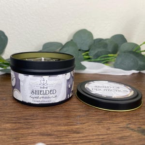 May include: A black tin candle with the text "SHIELDED" and "Shield of Protection" on the lid. The candle has a white label with a pattern of clocks and the scent is Citronella & Rosemary.