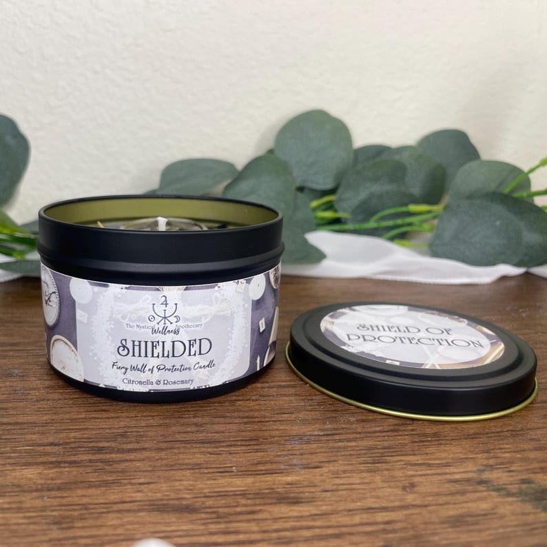 May include: A black tin candle with the text "SHIELDED" and "Shield of Protection" on the lid. The candle has a white label with a pattern of clocks and the scent is Citronella & Rosemary.