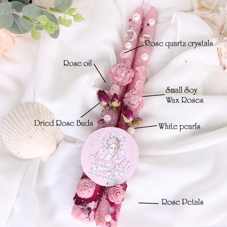 May include: Two pink candles decorated with rose quartz crystals, small soy wax roses, dried rose buds, white pearls, and rose petals. A round label with the word "Aphrodite" is placed on the candles. The candles are on a white surface.