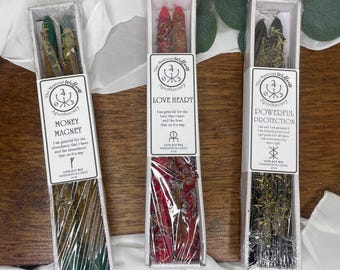 May include: Three boxed taper candles with labels reading "Money Magnet", "Love Heart", and "Powerful Protection". The candles are various colors, including green, red, and black, and are decorated with herbs. The packaging includes the brand name "The Mystical Wellness Apothecary".