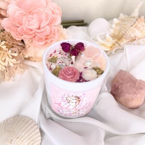 May include: A white candle with a pink label featuring the word "Aphrodite" and an illustration of a woman. The candle is decorated with pink roses, dried rosebuds, pearls, and gold-colored accents. A pink crystal and seashells are also visible.