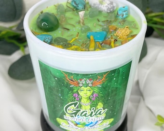 Könnte beinhalten: Eine weiße Kerze mit grünem Deckel, verziert mit verschiedenen Steinen und dekorativen Elementen. Die Kerze hat ein Etikett mit dem Wort "Gaia" und einer Illustration einer Frau. Die Kerze steht auf einem schwarzen Sockel.
