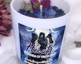 May include: A white candle with blue wax, decorated with dried flowers, black stones, and a charm. The label features the word "Hecate" and the text "Goddess of Witchcraft". The candle is made with lavender, rosemary, and black amber soy wax.