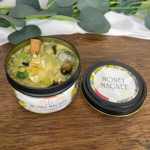 May include: A Money Magnet handmade crystal candle in a black tin. The candle is filled with green crystals, gold flakes, and a cinnamon stick. The lid, placed to the side, has the words "MONEY MAGNET" in white.