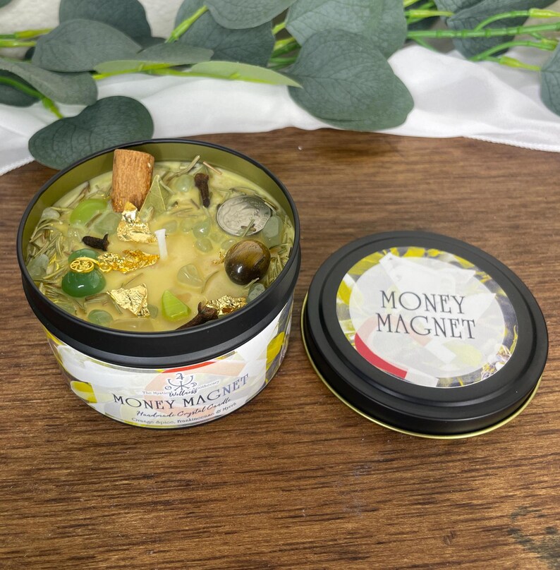 May include: A Money Magnet handmade crystal candle in a black tin. The candle is filled with green crystals, gold flakes, and a cinnamon stick. The lid, placed to the side, has the words "MONEY MAGNET" in white.