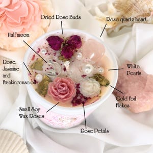 May include: A soy wax candle in a white container, adorned with dried rose buds, small wax roses, rose petals, white pearls, and gold foil flakes. A rose quartz heart and a half-moon charm add to the aesthetic. The candle is infused with rose, jasmine, and frankincense oil.