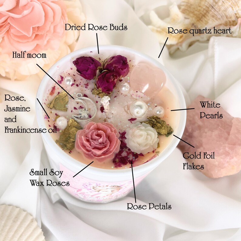 May include: A soy wax candle in a white container, adorned with dried rose buds, small wax roses, rose petals, white pearls, and gold foil flakes. A rose quartz heart and a half-moon charm add to the aesthetic. The candle is infused with rose, jasmine, and frankincense oil.