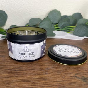 May include: A black tin candle with the word "SHIELDED" printed on the label. The label also includes the text "Fairy Wall of Protective Candle" and "Citronella & Rosemary". The lid is off, revealing the candle's contents and the words "SHIELD OF PROTECTION".