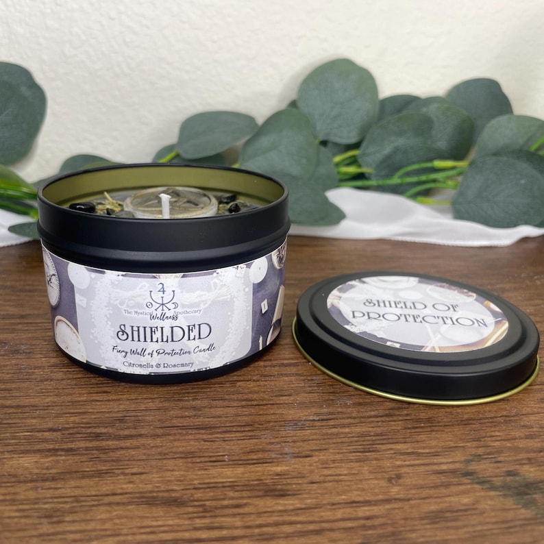 May include: A black tin candle with the word "SHIELDED" printed on the label. The label also includes the text "Fairy Wall of Protective Candle" and "Citronella & Rosemary". The lid is off, revealing the candle's contents and the words "SHIELD OF PROTECTION".