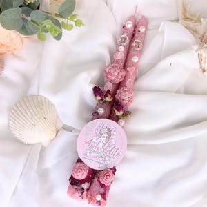 May include: Two pink, decorative candles adorned with pearls, dried roses, and crystals. The candles have a glittery finish and are embellished with small silver charms. A round sticker with the word "Aphrodite" and a woman's face is placed on the candles.
