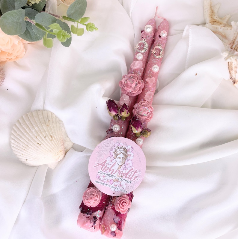 May include: Two pink, decorative candles adorned with pearls, dried roses, and crystals. The candles have a glittery finish and are embellished with small silver charms. A round sticker with the word "Aphrodite" and a woman's face is placed on the candles.