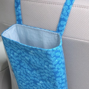 May include: A blue fabric trash bag with a white leaf pattern hangs from the headrest of a car seat. The bag is designed to hold trash in a vehicle.