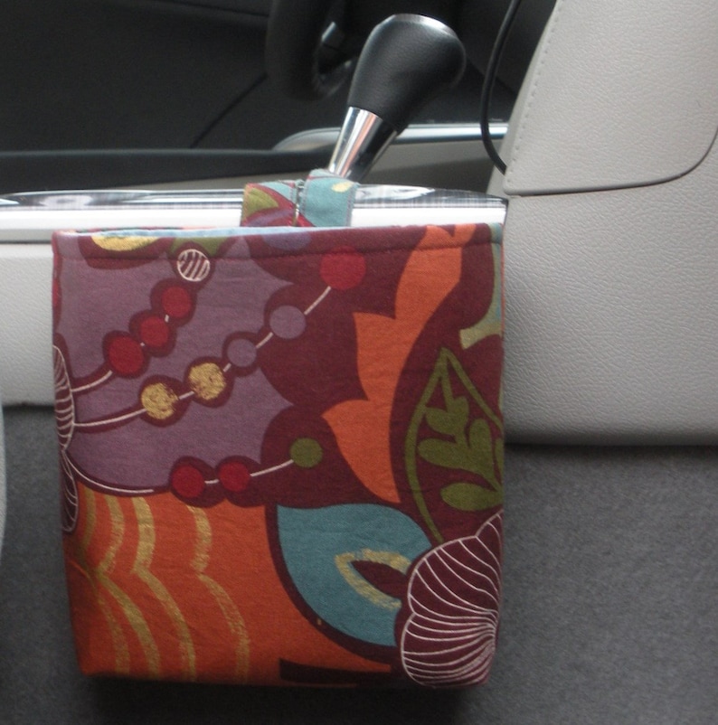 May include: A colorful fabric car organizer with a floral pattern in shades of orange, red, purple, and green. The organizer is hanging from the gear shift lever in a car.