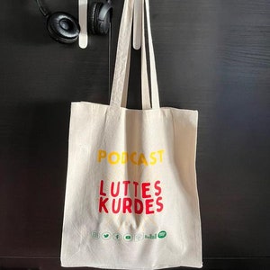 May include: A white canvas tote bag with the text "Podcast Luttes Kurdes" in yellow and red lettering. The bag has social media icons printed on the bottom.