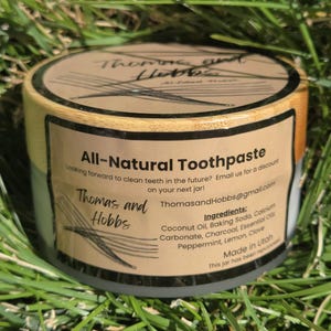 May include: A close-up of a brown and gray jar of all-natural toothpaste labeled "Thomas and Hobbs". The jar is sitting on a bed of green grass. The label lists the ingredients: Coconut Oil, Baking Soda, Calcium Carbonate, Charcoal, Essential Oils, Peppermint, Lemon, Clove. The jar is made in Utah.