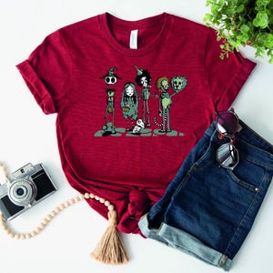 May include: Red t-shirt with a graphic print of a group of cartoon characters. The characters are a witch, a girl, a tin man, a lion, and a dog. The characters are all wearing different outfits and have different expressions on their faces.