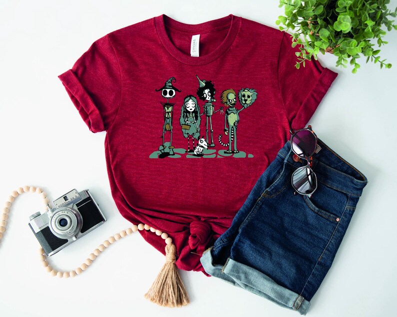 May include: Red t-shirt with a graphic print of a group of cartoon characters. The characters are a witch, a girl, a tin man, a lion, and a dog. The characters are all wearing different outfits and have different expressions on their faces.